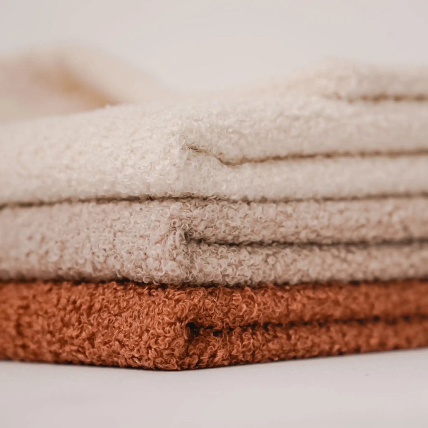 Zbliżenie na detale. 3 kocyki dla psa, w kolorze nugat (rudo-brązowy), pralinka (jasno brązowy), chałwa (kremowy), ułożone jeden na drugim od najjaśniejszego do najciemniejszego. Nowoczesna, wysokiej jakości tkanina boucle z certyfikatami bezpieczeństwa.