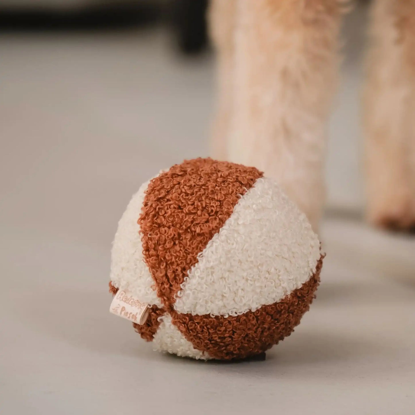 Piłka dla psa z tkaniny boucle. Kolory: rudo brązowy oraz jasny kremowy, piłka znajduje sie w salonie obok łapek pieska, bliski detal.