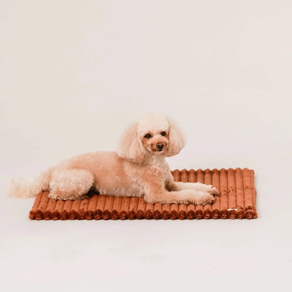 Piesek leży na puszystym kocyku dla psa, w kolorze nugat (rudo-brązowy). Kocyk ma falowaną strukturę. Jasne, czyste tło.