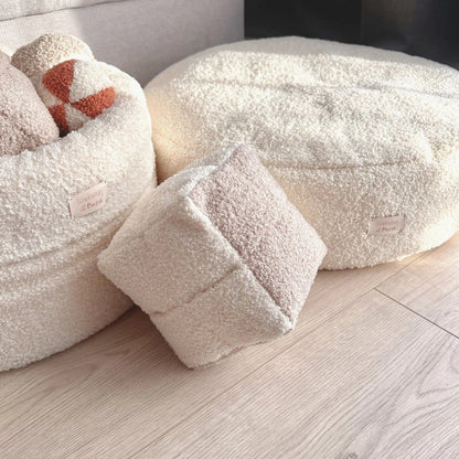 Miękka zabawka węchowa dla psa z tkaniny boucle w kształcie kostki do gry w kolorze chałwa-pralinka (kremowo-bezowa). Kostka na tle legowiska oraz kosza na zabawki dla psa, jasna, spójna kolorystyka.