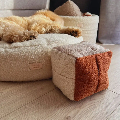 Miękka zabawka węchowa dla psa z tkaniny boucle w kształcie kostki do gry w kolorze chałwa-pralinka-nugat (kremowy-bezowy-ceglasty). Kostka na tle pieska leżącego na legowisku oraz kosza na zabawki dla psa, jasna, spójna kolorystyka. Ujęcie z od dołu.