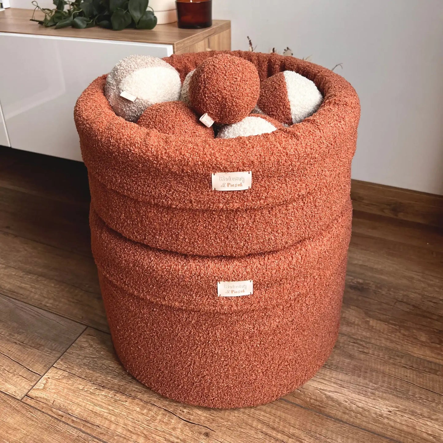 2 kosze na zabawki dla psa, tkanina boucle, kolor nugat (terakota), kosz znajduje się w jasnym wnętrzu, wypełniony jest piłeczkami z tej samej tkaniny w różnych kolorach, porównanie dwóch rozmiarów, niższego i wyższego, kosze ułożone na sobie