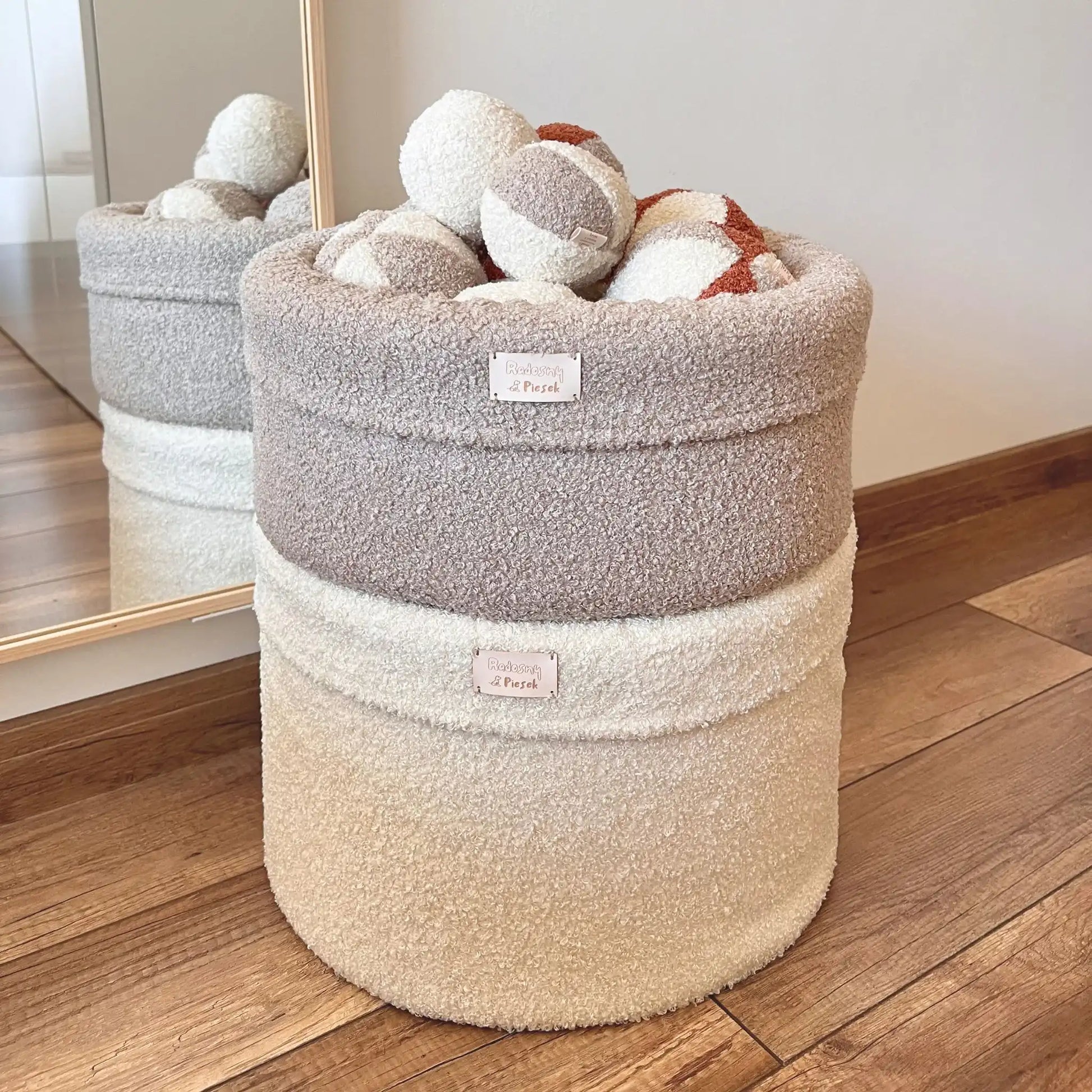 2 Miękkie kosze na zabawki dla psa, wypełnione pianką tapicerską, uszyte z tkaniny boucle w kolorze pralinka (jasny brąz) oraz chałwa (kremowy). Kosze ułożone są na sobie, większy rozmiar na dole, mniejszy rozmiar na górze. Górny kosz wypełniony piłeczkami w kolorach kremowo brązowych. Jasne wnętrze.