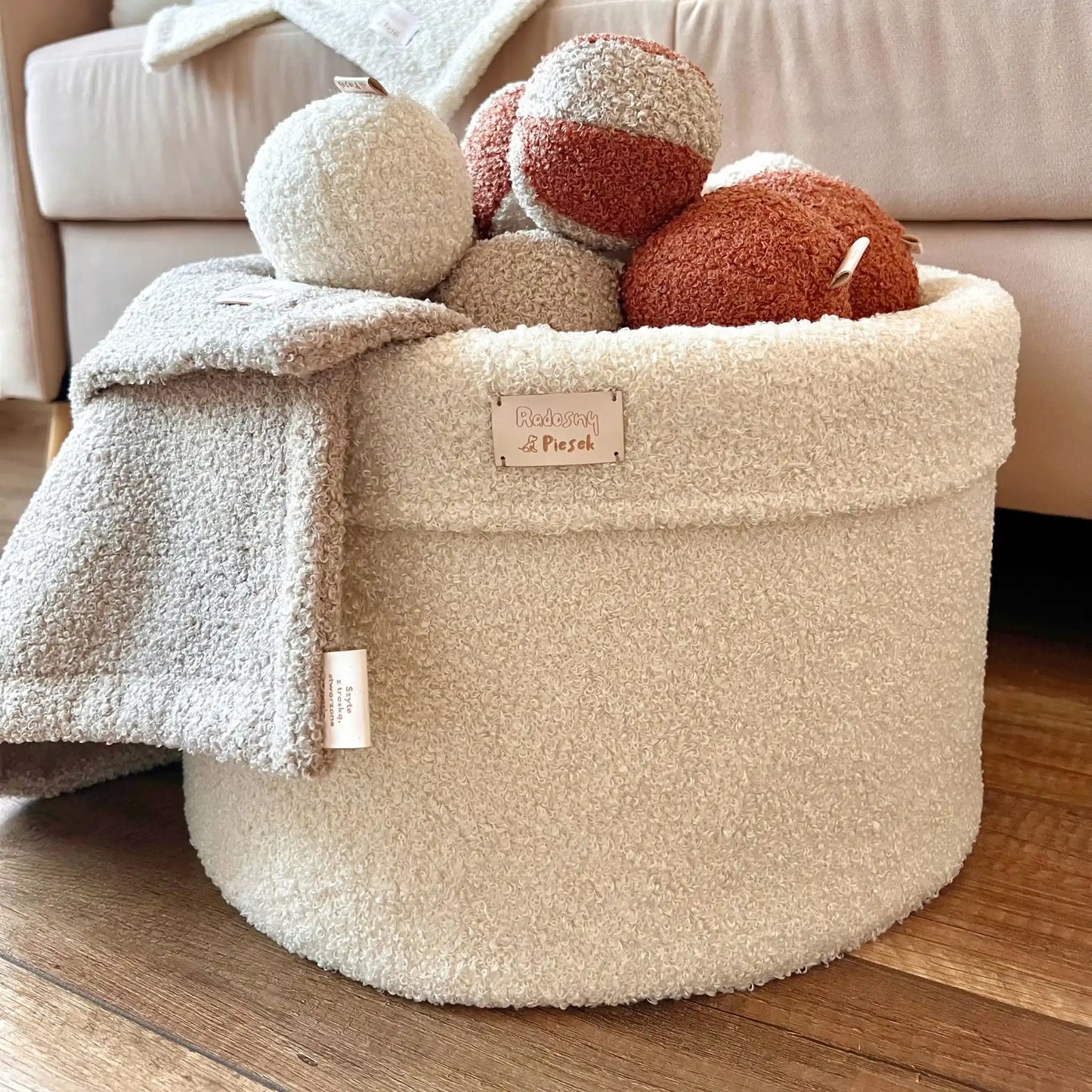 Miękki kosz na zabawki dla psa. Usztywniony pianką tapicerską. Uszyty z tkaniny boucle w kolorze chałwa (kremowy) wypełniony piłeczkami w kolorach kremowo brązowych. Kosz stoi obok kanapy w salonie. Zbliżenie.