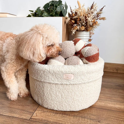 Miękki kosz na zabawki dla psa. Usztywniony pianką tapicerską. Uszyty z tkaniny boucle w kolorze chałwa (kremowy) wypełniony piłeczkami w kolorach kremowo brązowych. Kosz stoi obok szafki rtv w salonie, piesek - pudel miniturowy zagląda do środka. 