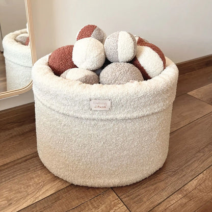 Miękki kosz na zabawki dla psa. Usztywniony pianką tapicerską. Uszyty z tkaniny boucle w kolorze chałwa (kremowy) wypełniony piłeczkami w kolorach kremowo brązowych. Kosz stoi na podłodze obok lustra.