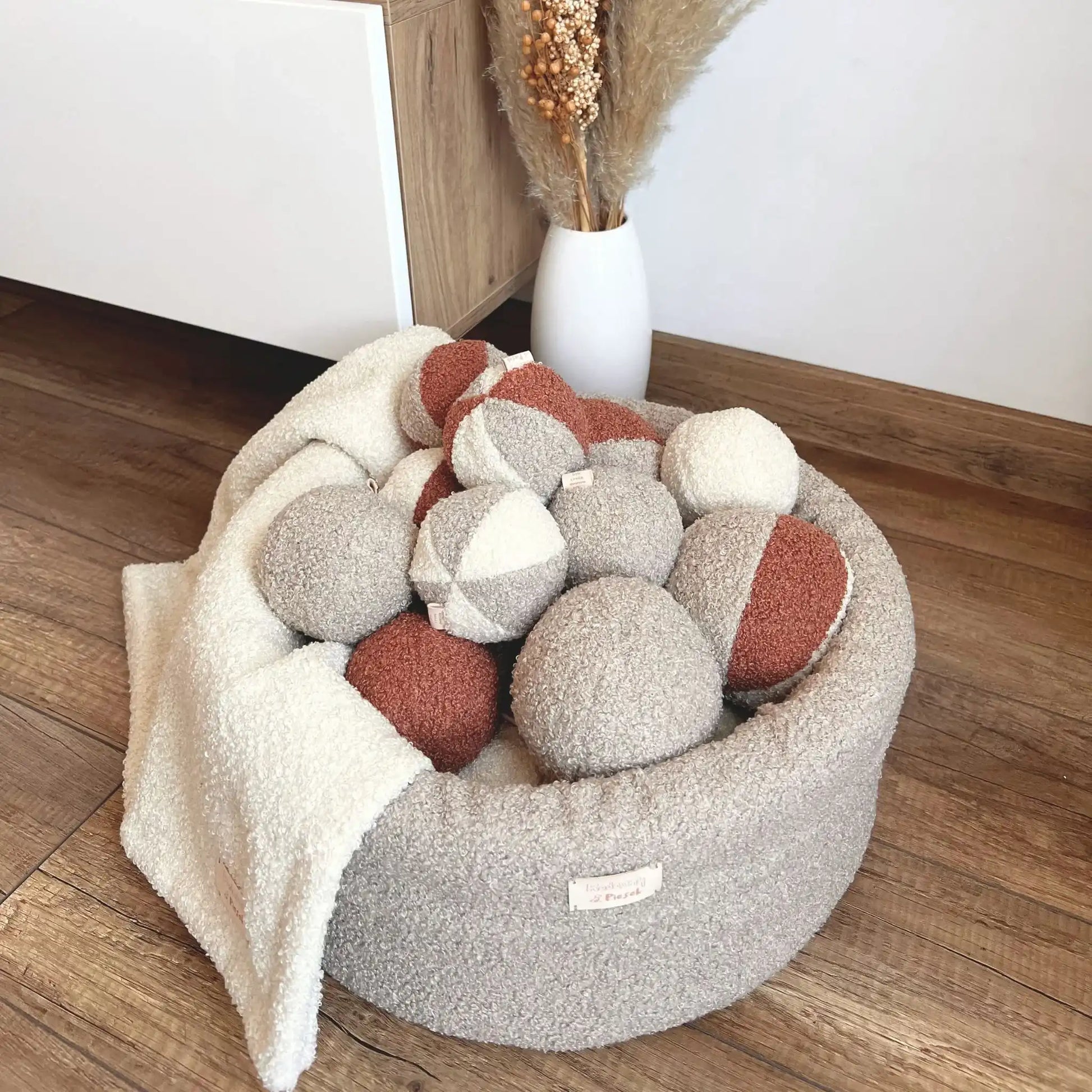 Miękki kosz na zabawki dla psa. Usztywniony pianką tapicerską. Uszyty z tkaniny boucle w kolorze pralinka (jasny brąz) wypełniony piłeczkami w kolorach kremowo brązowych. Kosz stoi obok szafki rtv w salonie.