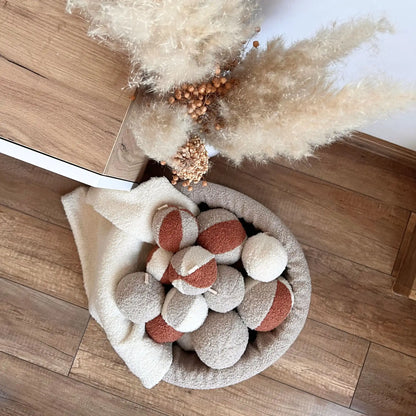 Miękki kosz na zabawki dla psa. Usztywniony pianką tapicerską. Uszyty z tkaniny boucle w kolorze pralinka (jasny brąz) wypełniony piłeczkami w kolorach kremowo brązowych. Kosz stoi obok szafki rtv w salonie. Ujęcie z góry. Obok stoi nowoczesna kompozycja suszonych traw.