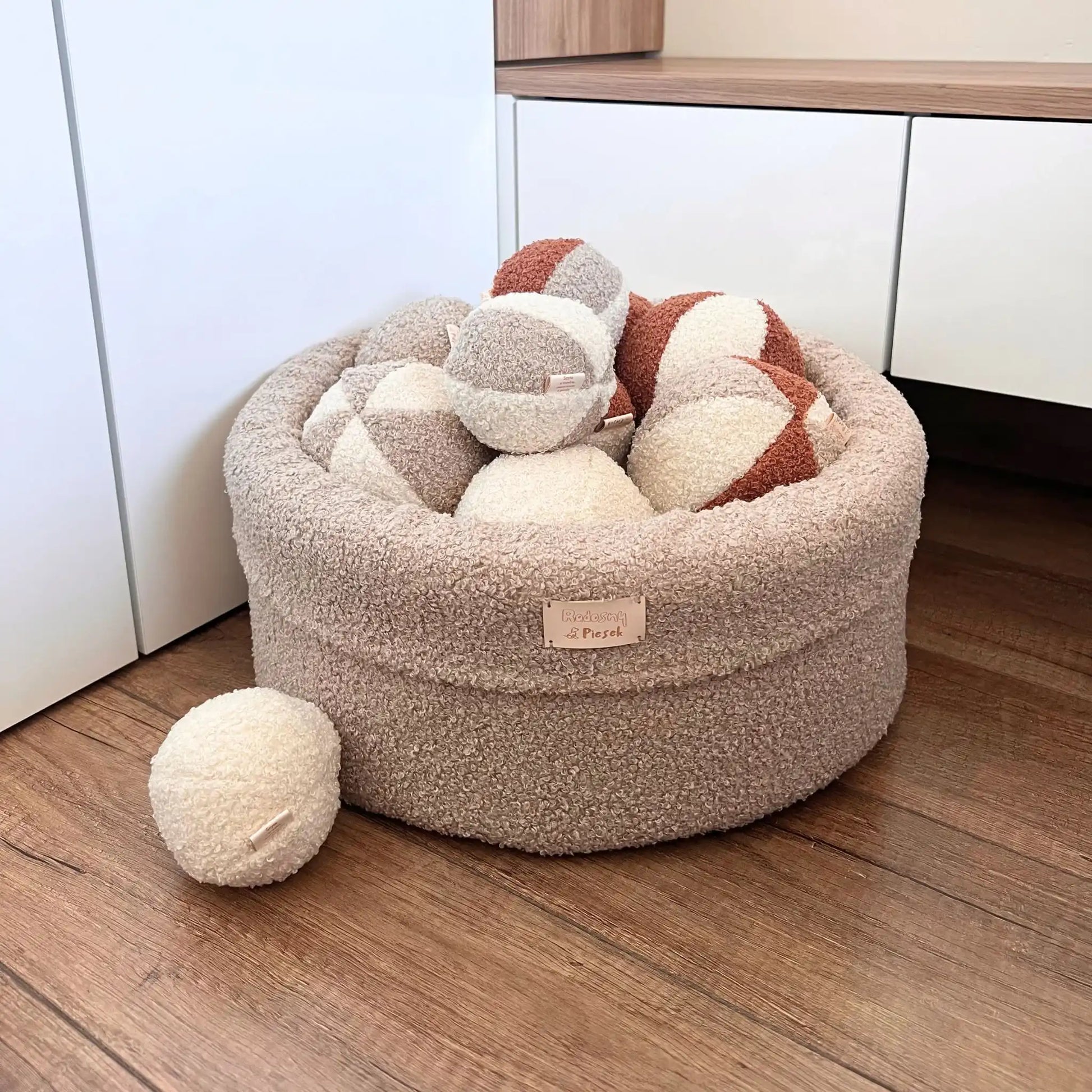 Miękki kosz na zabawki dla psa. Usztywniony pianką tapicerską. Uszyty z tkaniny boucle w kolorze pralinka (jasny brąz) wypełniony piłeczkami w kolorach kremowo brązowych. Kosz stoi obok szafki nocnej w sypialni w jasny wnętrzu.