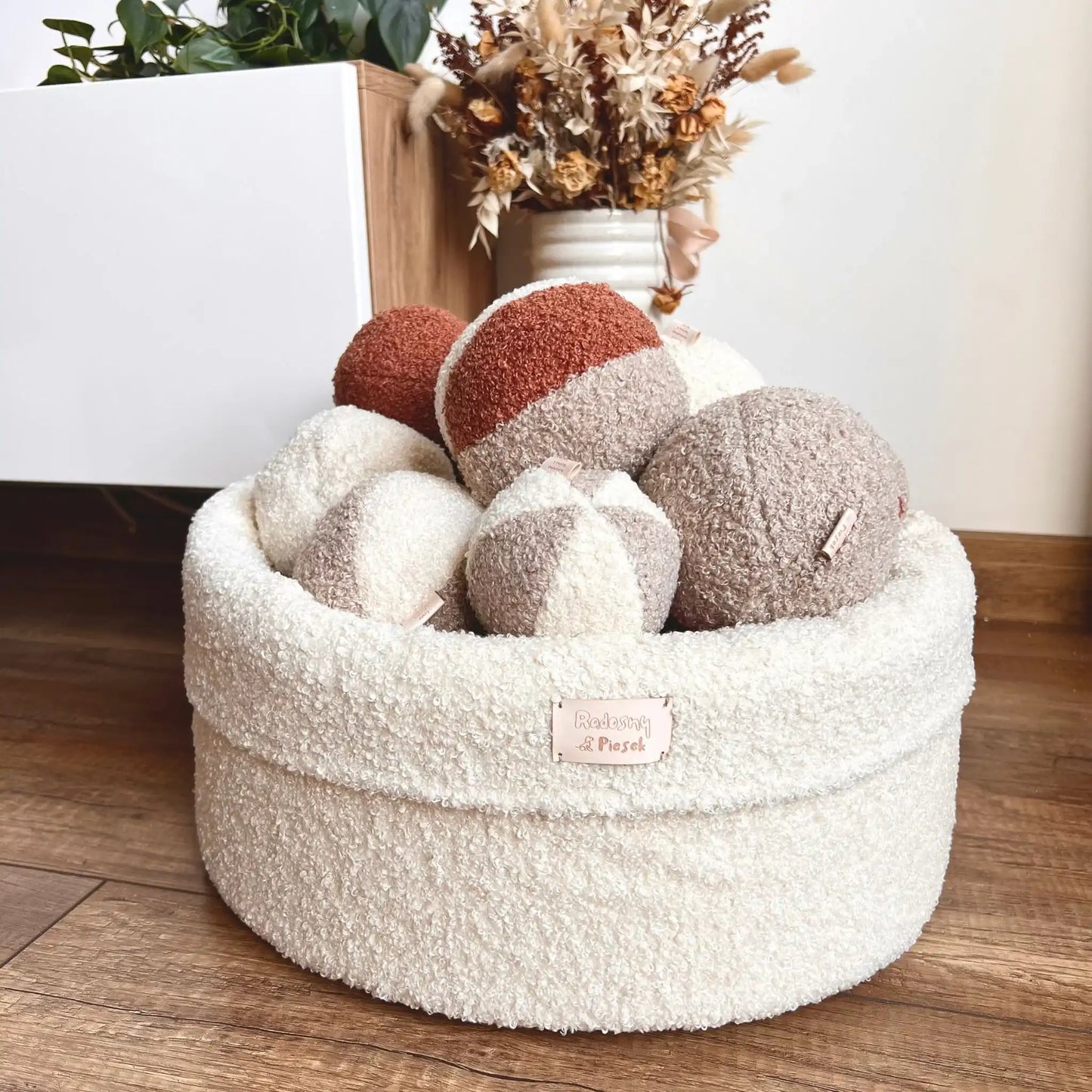 Miękki kosz na zabawki dla psa. Usztywniony pianką tapicerską. Uszyty z tkaniny boucle w kolorze chałwa (kremowy) wypełniony piłeczkami w kolorach kremowo brązowych. Kosz stoi obok szafki rtv w salonie.