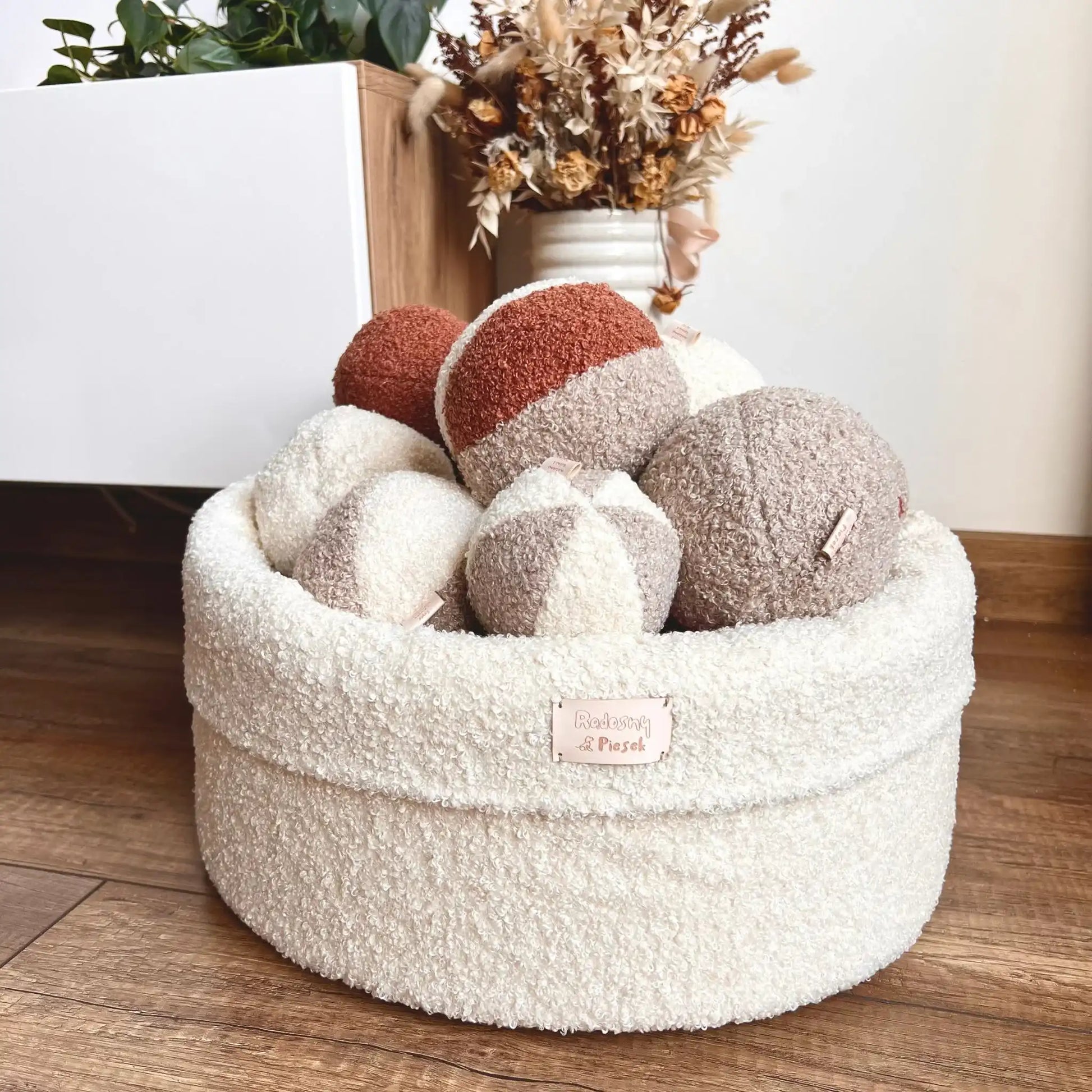 Miękki kosz na zabawki dla psa. Usztywniony pianką tapicerską. Uszyty z tkaniny boucle w kolorze chałwa (kremowy) wypełniony piłeczkami w kolorach kremowo brązowych. Kosz stoi obok szafki rtv w salonie.