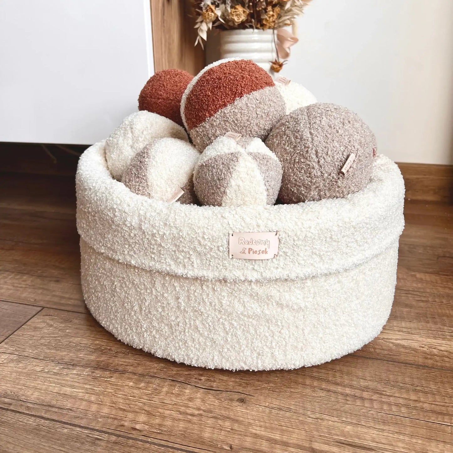 Miękki kosz na zabawki dla psa. Usztywniony pianką tapicerską. Uszyty z tkaniny boucle w kolorze chałwa (kremowy) wypełniony piłeczkami w kolorach kremowo brązowych. Kosz stoi obok szafki rtv w salonie.