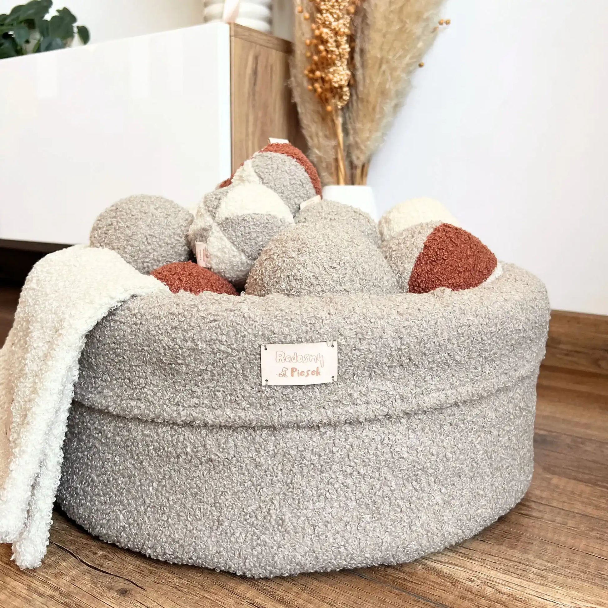 Miękki kosz na zabawki dla psa. Usztywniony pianką tapicerską. Uszyty z tkaniny boucle w kolorze pralinka (jasny brąz) wypełniony piłeczkami w kolorach kremowo brązowych. Kosz stoi obok szafki rtv w salonie.