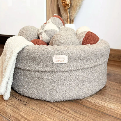 Miękki kosz na zabawki dla psa. Usztywniony pianką tapicerską. Uszyty z tkaniny boucle w kolorze pralinka (jasny brąz) wypełniony piłeczkami w kolorach kremowo brązowych. Kosz stoi obok szafki rtv w salonie.