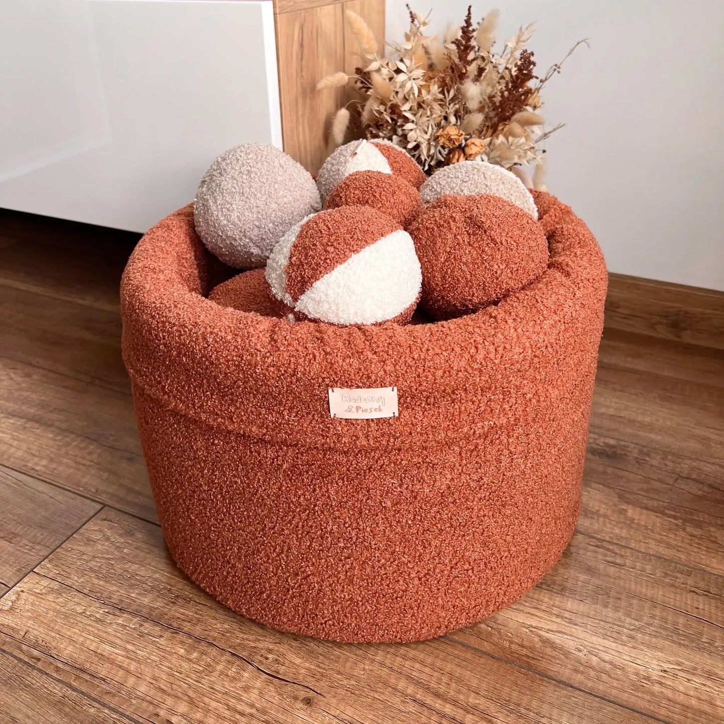 kosz na zabawki dla psa, tkanina boucle, kolor nugat (terakota), kosz znajduje się w jasnym wnętrzu, wypełniony jest piłeczkami z tej samej tkaniny w różnych kolorach