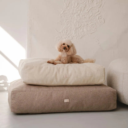 Piesek leży na dwóch (ułożonych na sobie w rozmiarze m i l) miękkich legowiskach dla psa (materacach) w kolorze pralinka (jasny brąz) oraz chałwa (kremowy), które znajdują się w jasnym, nowoczesnym wnętrzu na tle kanapy oraz nowoczesnego obrazu w jednolitym kolorze). Nowoczesna, wysokiej jakości tkanina boucle z certyfikatami bezpieczeństwa.