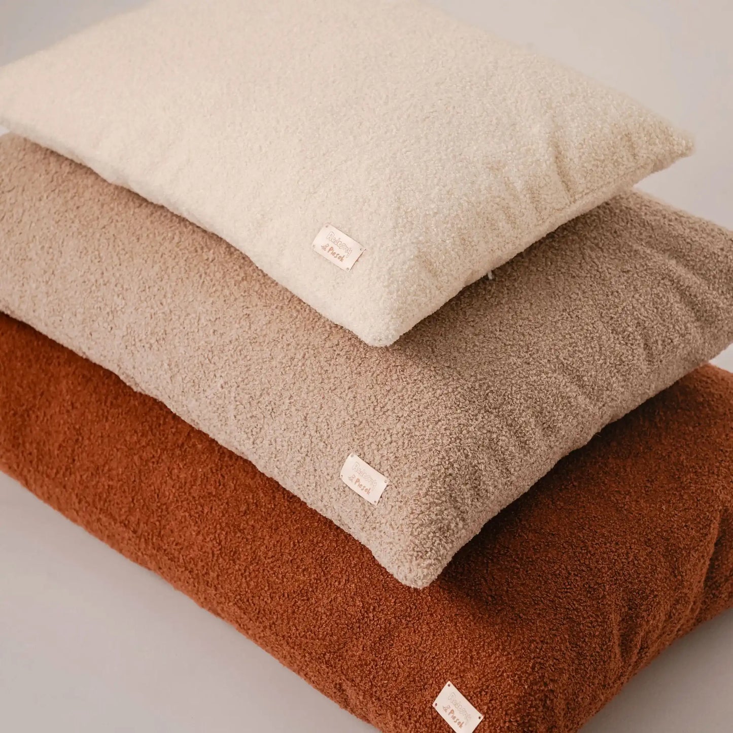 Zbliżenie na trzy poduszki dla psa (legowiska dla psa), w trzech rozmiarach, od największego do najmniejszego, w kolorze nugat (rudy brąz), pralinka (jasny brąz), chałwa (kremowy), które znajdują się na jasnym czystym tle. Nowoczesna, wysokiej jakości tkanina boucle z certyfikatami bezpieczeństwa.