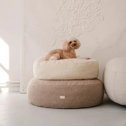 Piesek leży na dwóch (ułożonych na sobie w rozmiarze m i l) miękkich legowiskach dla psa (pufa dla psa) w kolorze pralinka (jasny brąz) oraz chałwa (kremowy), które znajdują się w jasnym, nowoczesnym wnętrzu na tle kanapy oraz nowoczesnego obrazu w jednolitym kolorze). Nowoczesna, wysokiej jakości tkanina boucle z certyfikatami bezpieczeństwa.