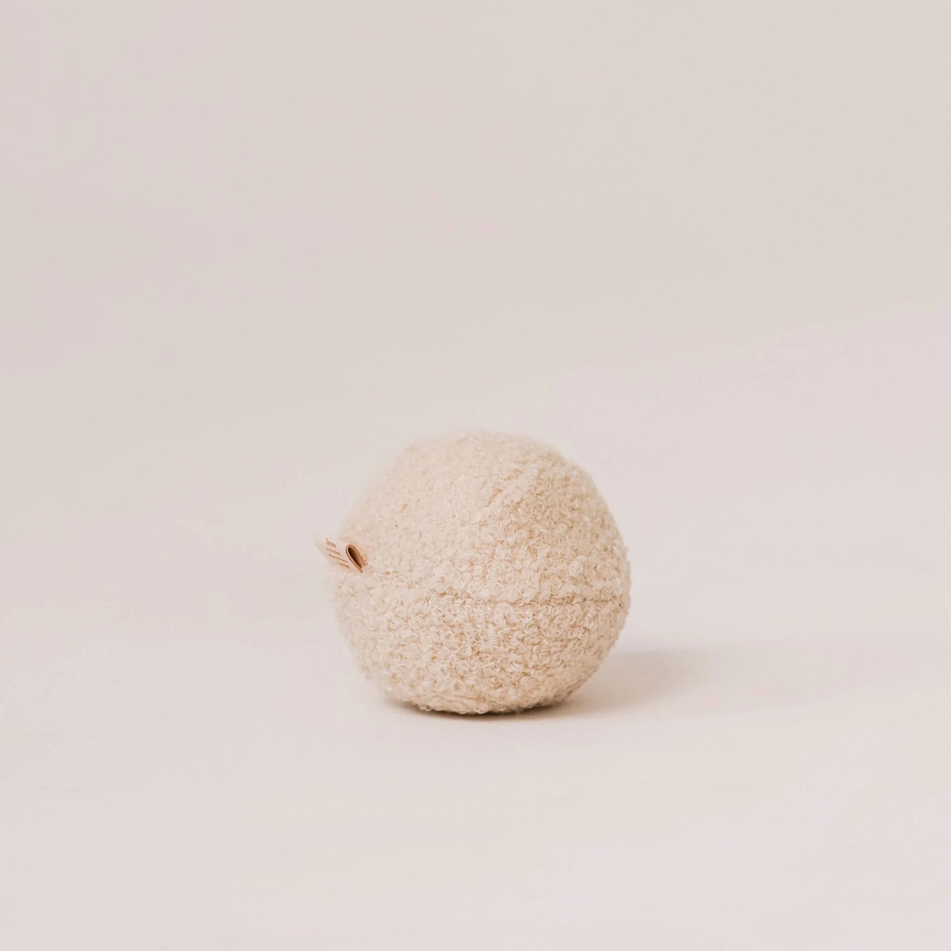 Miękka piłka dla psa z tkaniny boucle w kolorze chałwa (kremowy). Widoczna metka z logo. Jasne, czyste tło.