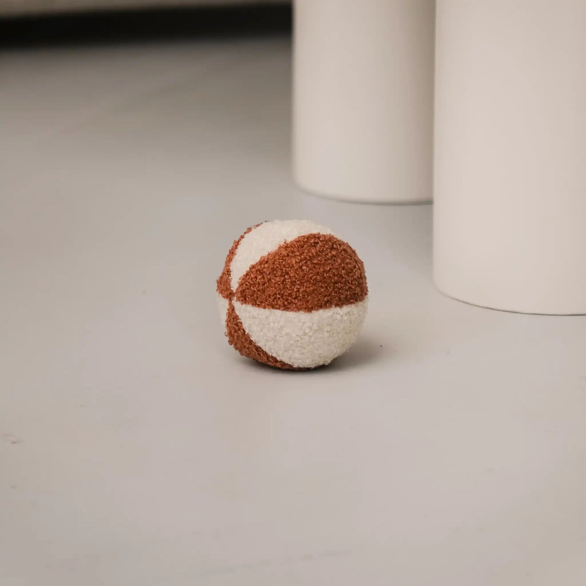 Piłka dla psa z tkaniny boucle. Kolory: rudo brązowy oraz jasny kremowy, piłka znajduje sie w salonie obok stołu, bliski detal.